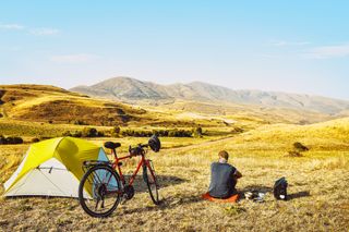 A cyclisr looks over a landscape with his bike and tent next to him