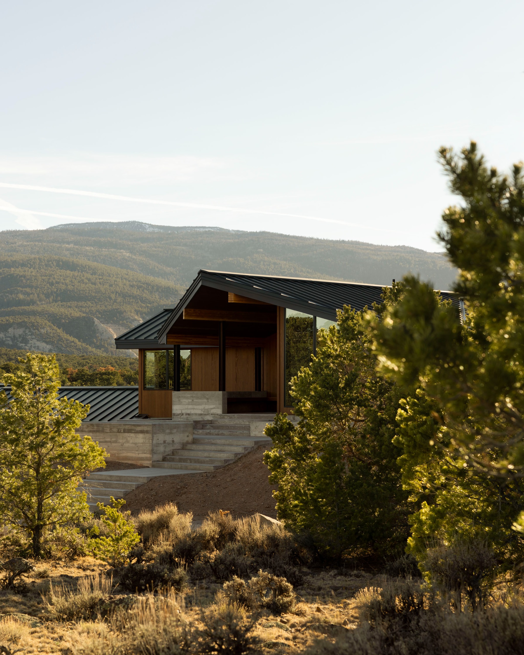 A view of a modern Utah desert home in the middle of the countryside with trees and foliage all around, mountainous areas in the background and a sunny clear sky