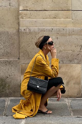 Spring trench coats shown in a photo of a woman crouching on the sidewalk in Paris wearing a black headband, black sunglasses, gold earrings, a yellow suede trench coat, black top, black capri pants, black heeled mules, and a black flap quilted shoulder bag