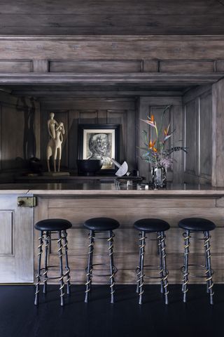 A wood bar with four brown stools, a small statute in the backdrop with a painting and a floral display.