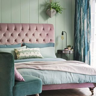 Bedroom with padded pink velvet bedstead, light blue velvet chair and cotton bedding in pastel shades on pale green panelled walls