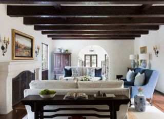 traditional living room with blue and white sofas dark ceiling beams stone fireplace with gilt framed picture above and dark wood console and cast iron details in background