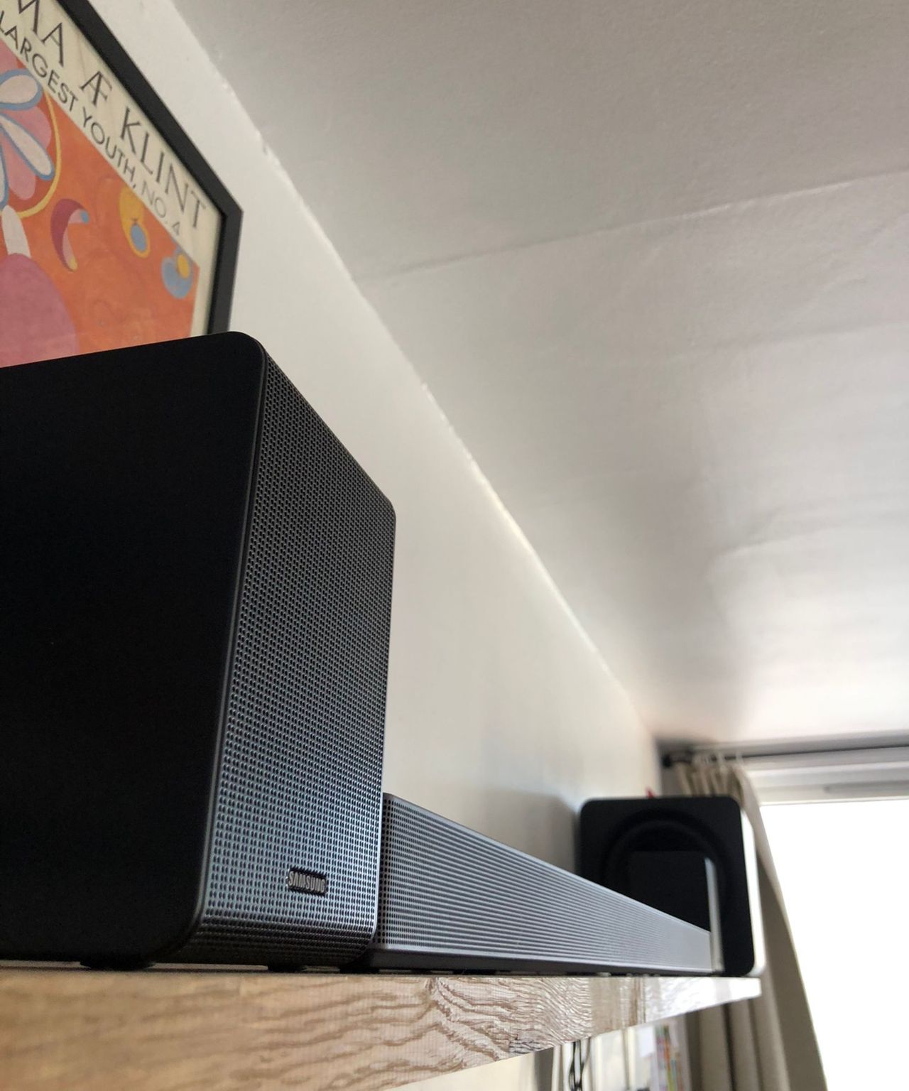 Samsung HW-QS750F soundbar on wooden shelf in Dan Fauzi's home, with curtains and bright window in background and art print pictured to the left.