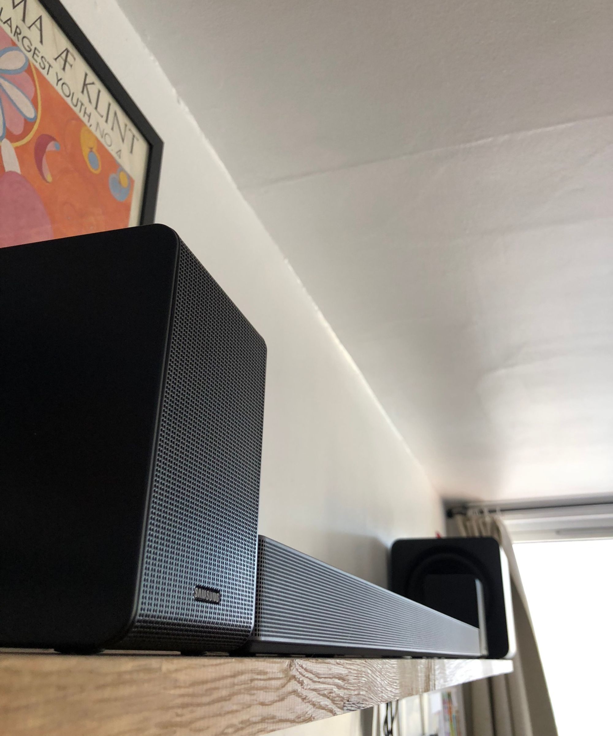 Samsung HW-QS750F soundbar on wooden shelf in Dan Fauzi&#039;s home, with curtains and bright window in background and art print pictured to the left.