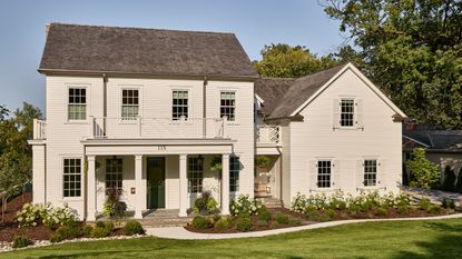 A large home exterior with warm white timber-clad walls, a front porch with four pillars, traditional-style windows and a dark green front door. A garden at the front of the house with lush green grass and a curved path.