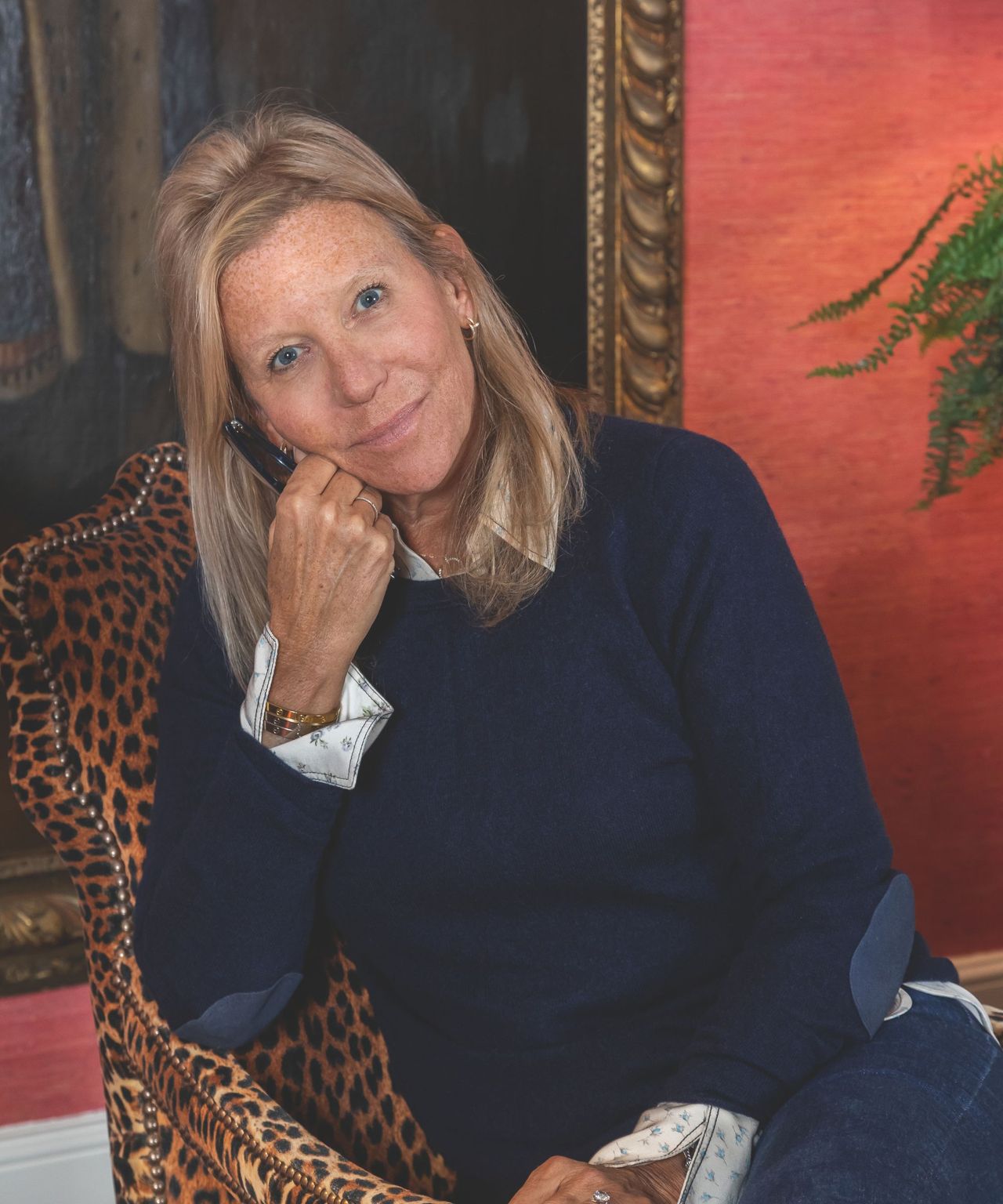 A headshot of a woman with shoulder length blond hair, wearing a navy jumper sits on a leopard print armchair