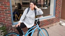 Woman with new bike, laughing and smiling on street outside a shop, wearing jumper and backpack