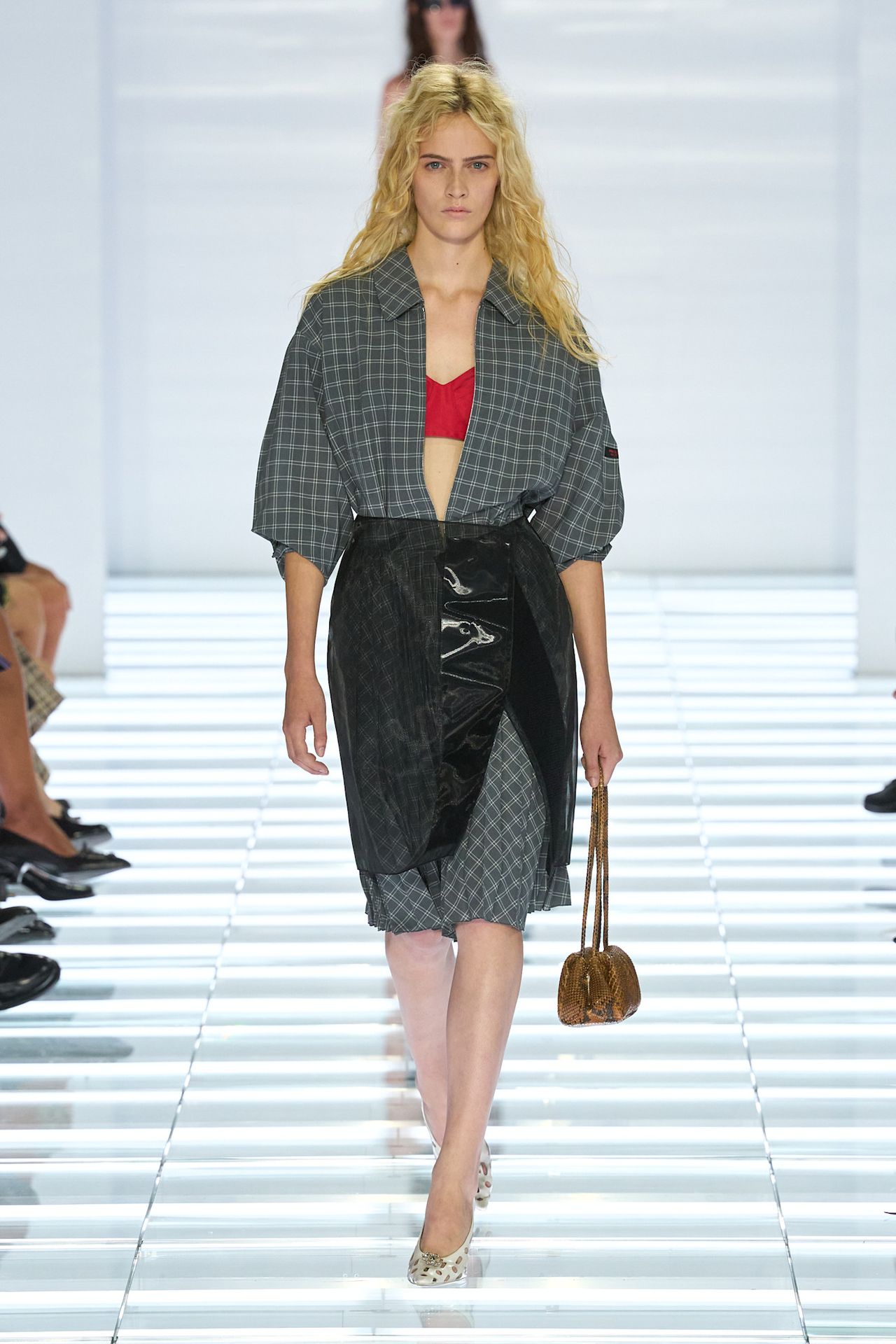 A Prada spring/summer 2026 runway model wearing a gray flannel dress layered under a sheer PVC black pencil skirt styled with heels.