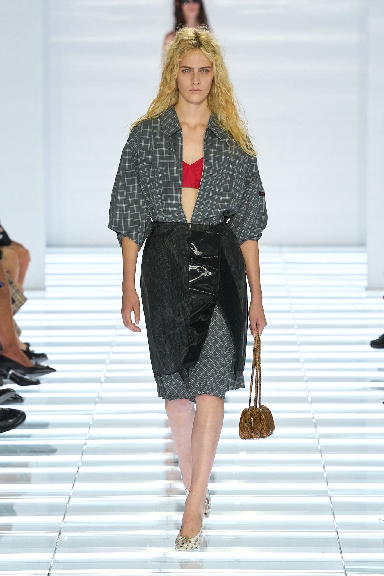 A Prada spring/summer 2026 runway model wearing a gray flannel dress layered under a sheer PVC black pencil skirt styled with heels.