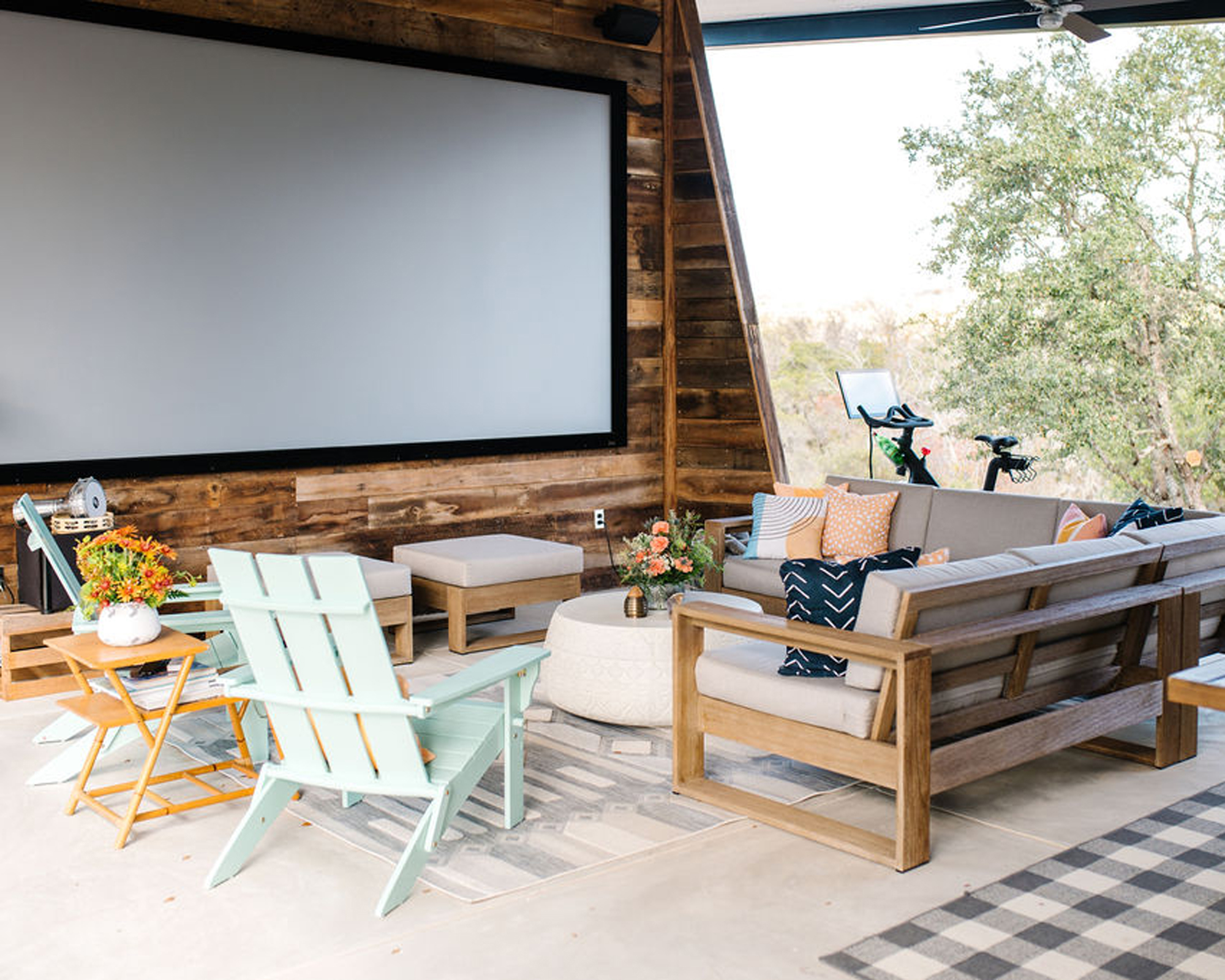 Rustic lounge with pastel pops via chairs and scatter pillows, and wood paneling on wall, with giant screen.