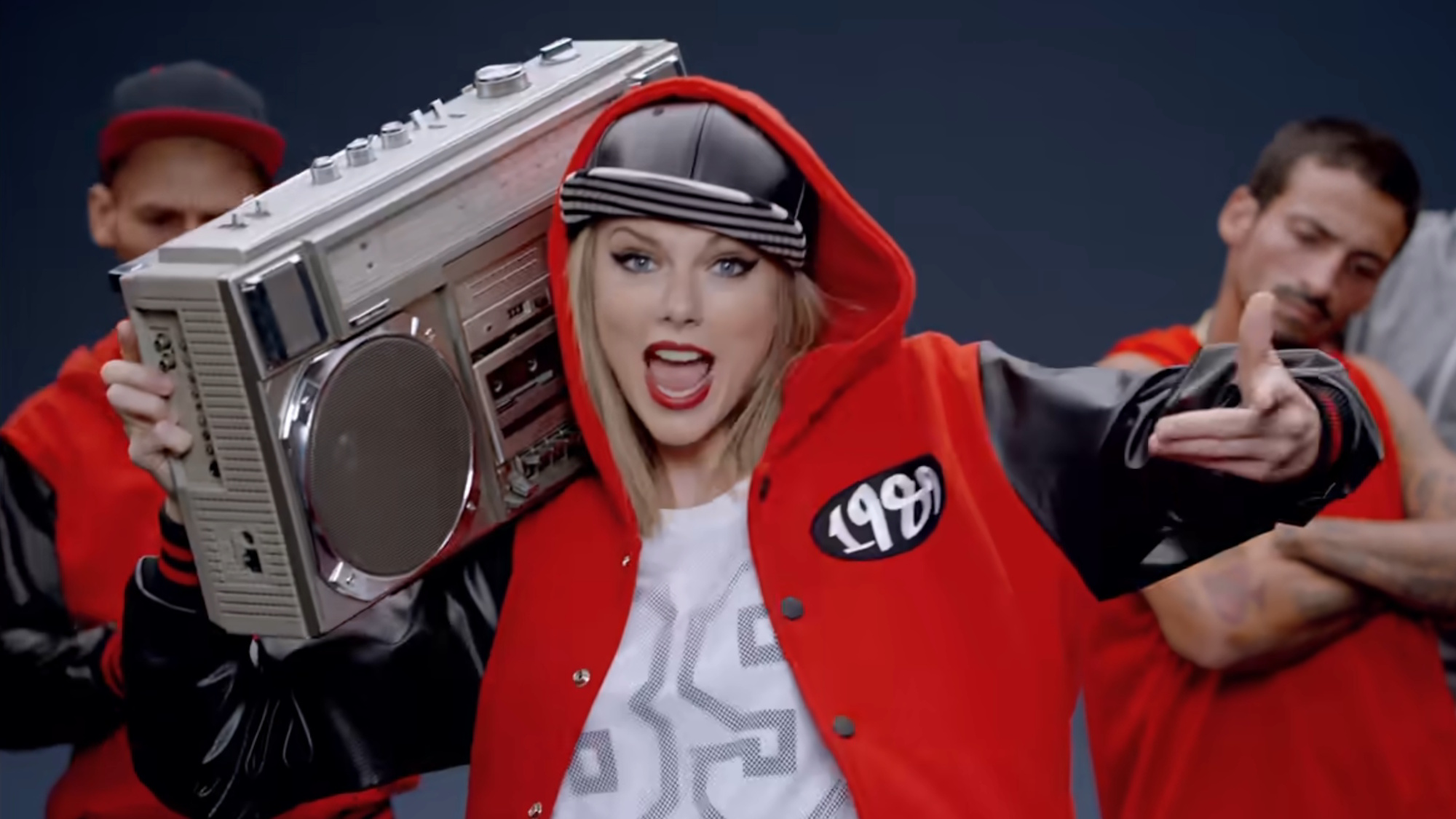 Taylor Swift is shown in the music video for Shake It Off wearing a white 89 tee with a red jacket and black cap, holding a boom box.