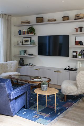 living room with tv on wall, blue couch, floating shelving, boucle armchairs, oval coffee table
