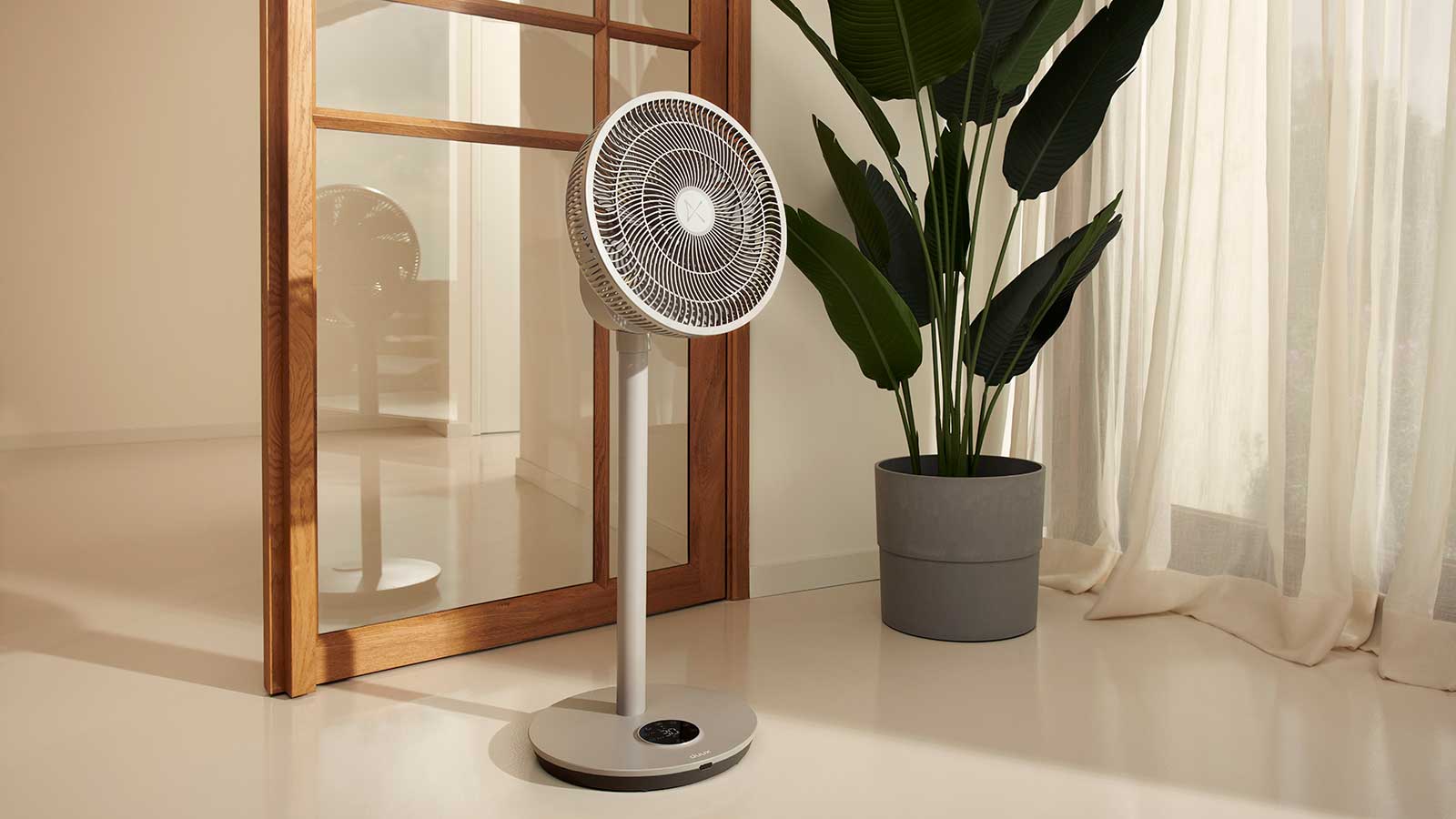 Duux Whisper Flex 2 pedestal fan in an off-white stone colour in a neutral room with a large houseplant behind the fan