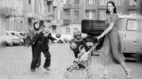 Black-and-white photo of a woman pushing a stroller past children playing with balls on a cobblestone street in a residential neighborhood