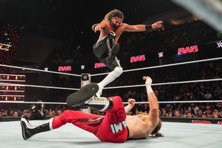 Seth Rollins is mid-air with his right foot outstretched, attempting a Curb Stomp move against Sami Zayn, who is lying on his back in the wrestling ring. The marquee screens in the background above the crowd show the "WWE" and "Raw" logos in repeat.