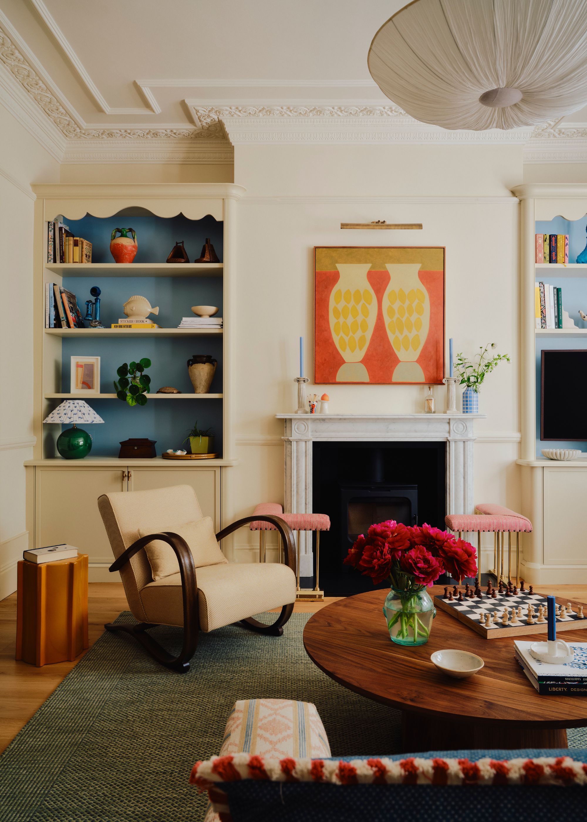 A living room in a period property with warm white walls and two alcoves with a sky blue backing. A fireplace with artwork above it, a round wood coffee table, a green rug, and a cream armchair.