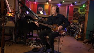 image of Sir Paul McCartney playing the guitar, creating Now and Then song. Behind him is Beatles memorabilia and guitars