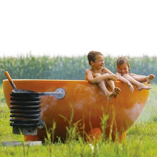 Two children sitting in a bowl-shaped hot tub outdoors with their legs dangling out over the side