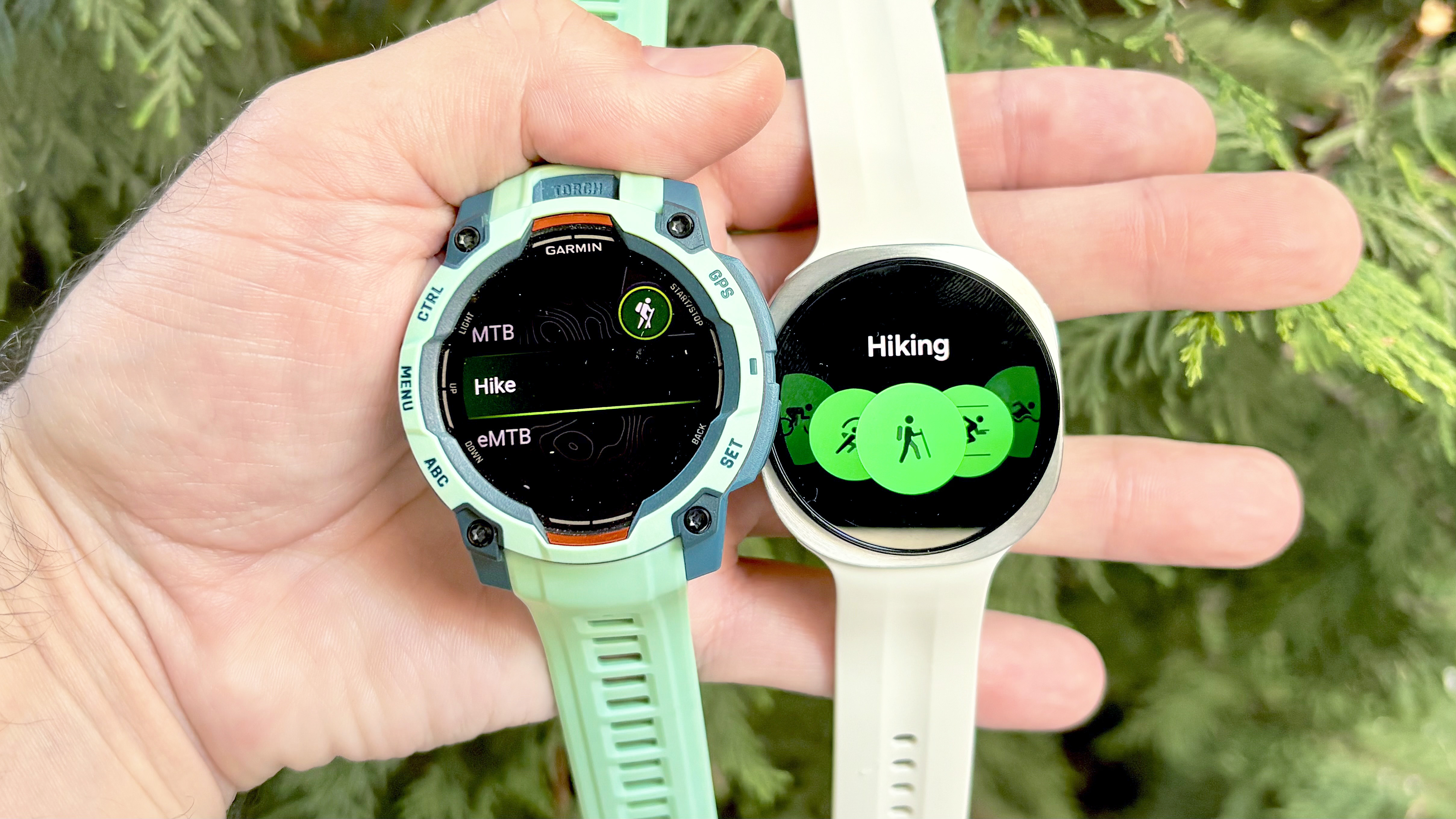 Close-up of the Garmin Instinct 3 next to the Samsung Galaxy Watch 9 with evergreens in the background
