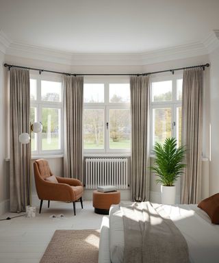beige curtain in bay window of large bright bedroom with orange armchair, houseplant and stool