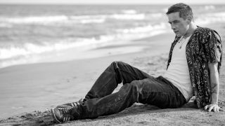 A black and white still from "Kaos", showing Orpheus (Killian Scott) sitting on a beach