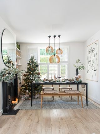 A neutral, open-plan living room with black dining room table, light wood bench, round mirror above black fireplace and trio of rattan ceiling lights