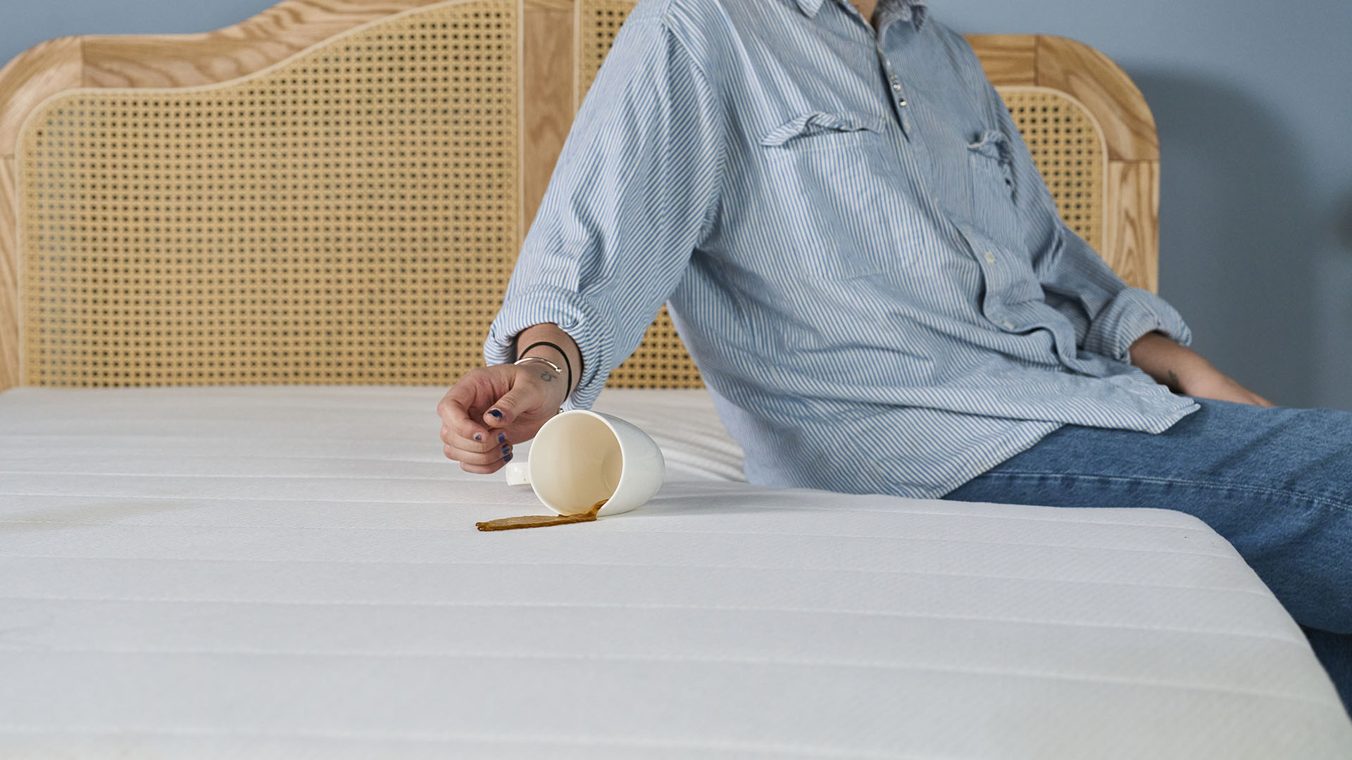 A close up of a person sitting on the edge of a mattress on a bed frame in a bedroom. Her arm is knocking over a white mug which has left a coffee stain on a mattress