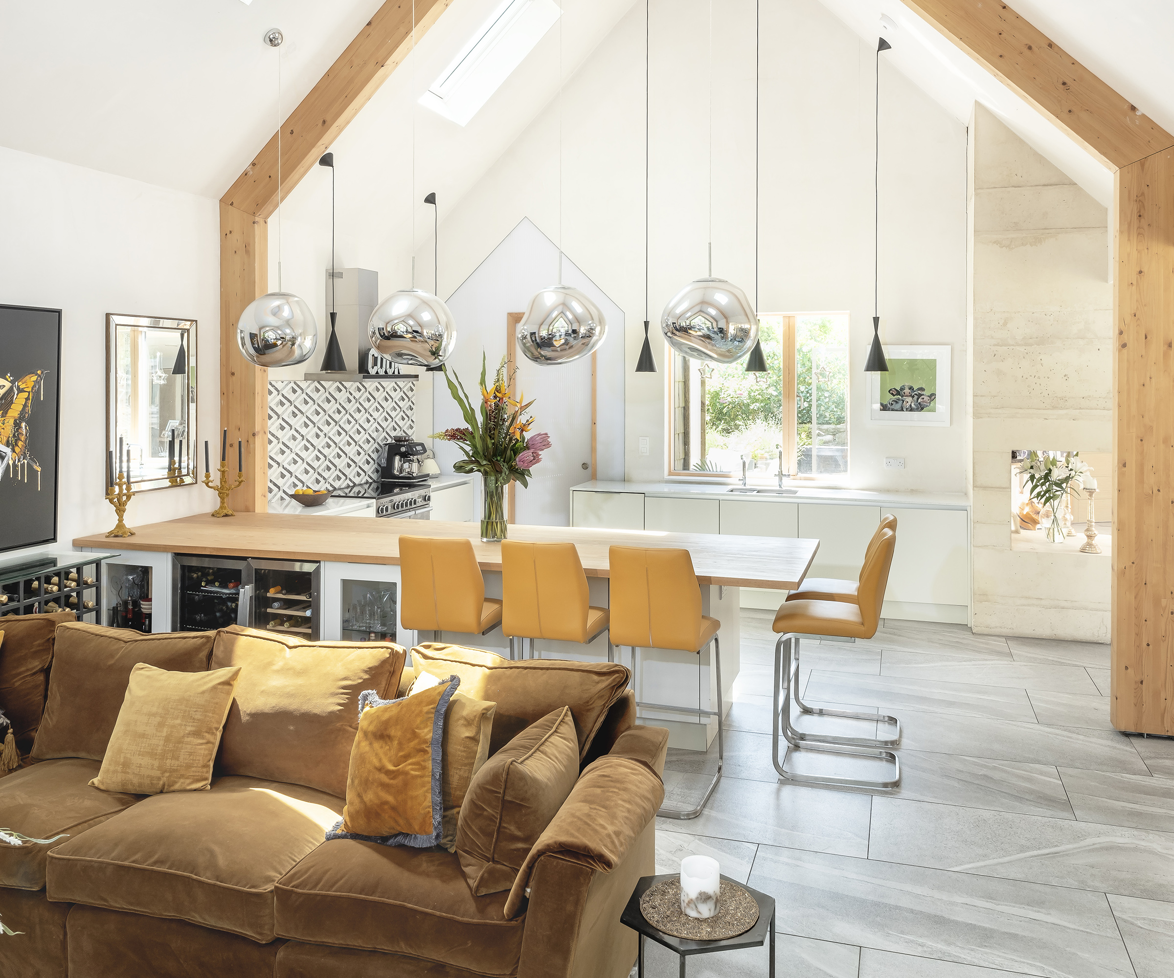 A kitchen with large peninsula with bar seating. The ceiling is pitched with exposed wooden beams. There are two styles of pendant light and lots of natural light from ceiling glazing