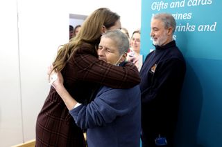 Kate Middleton wearing a burgundy plaid coat hugging a woman in a blue sweatshirt in front of a blue wall