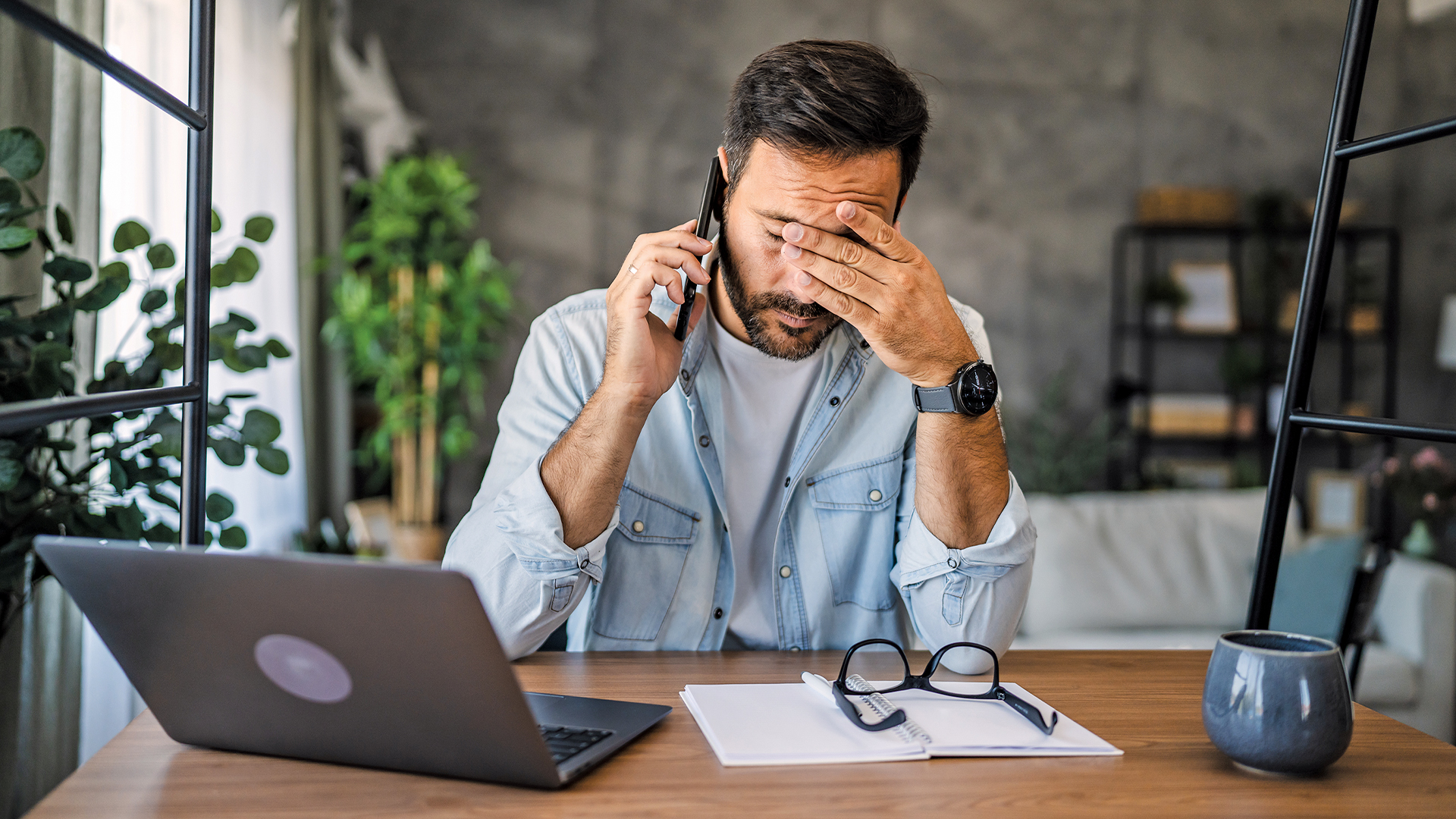 A man sits at the desk, a laptop in front of him, a phone in his hand, as he rubs his eyes tiredly in a worried expression