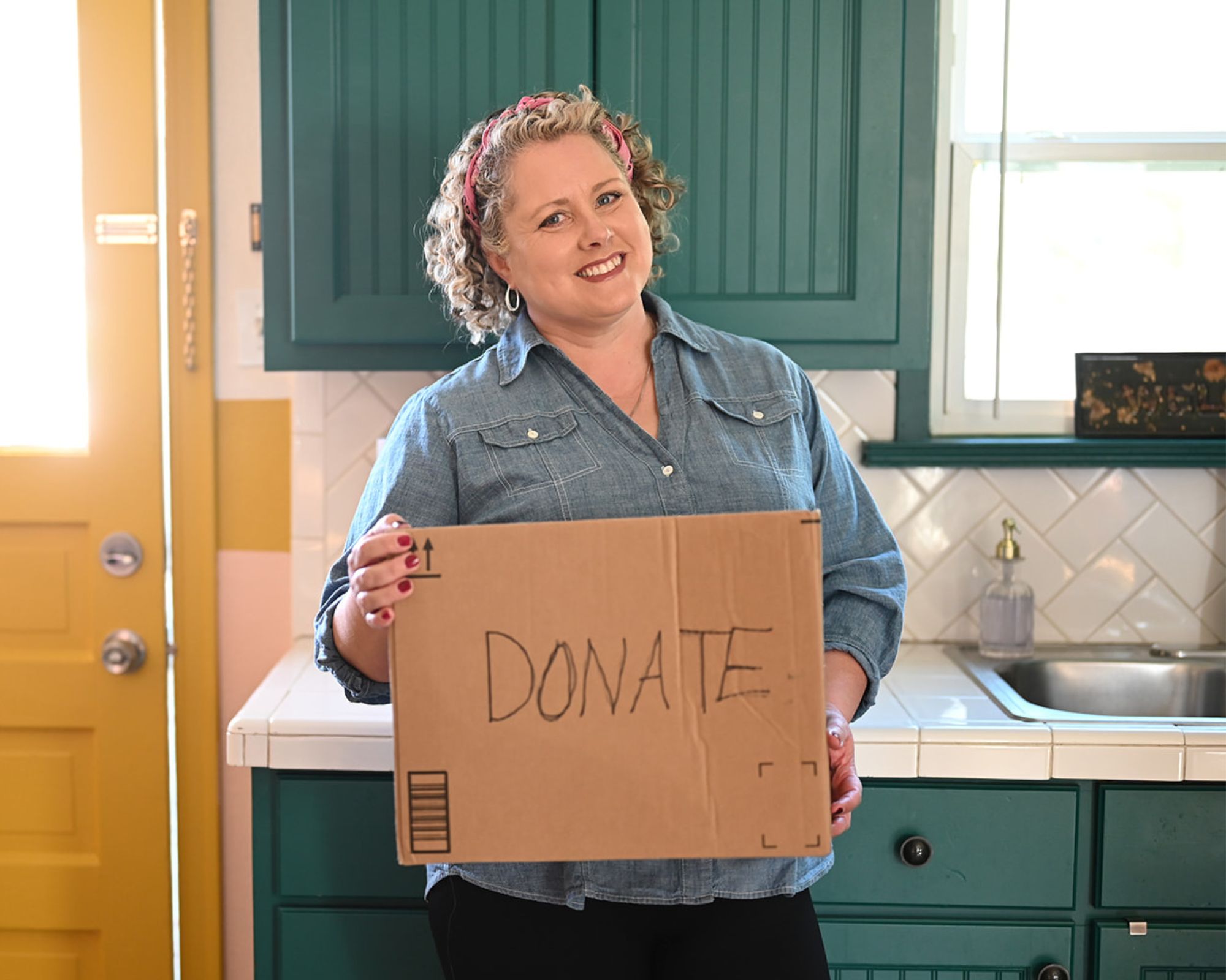 Dana White is a white woman with curly blonde hair. She's pictured wearing a denim shirt, holding a cardboard box with the word DONATE in black pen. She's in a green kitchen with tiled white subway backsplash and a yellow external door with bright light pouring in two windows