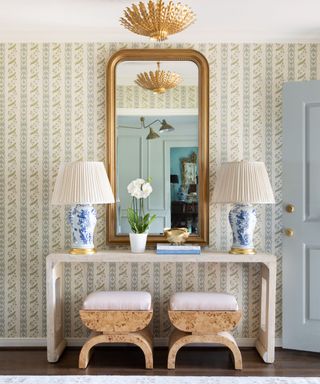 A soft blue entryway with wallpapered walls and a console with burl wooden stools and decorative lighting.