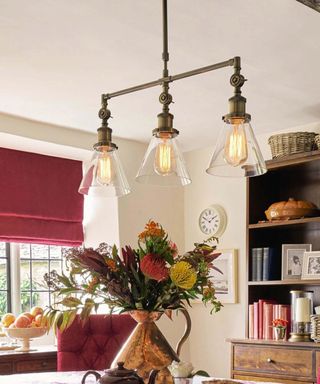 glass and brushed gold pendant light with three glass shades above copper jug with flowers, wooden dresser in background and dark red roman blind at window