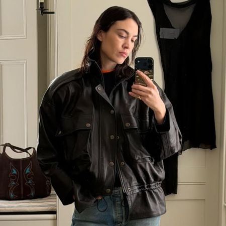 Alexa Chung takes a mirror selfie wearings straight-leg jeans with loafers and a cinch waist leather jacket.