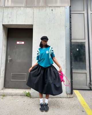 Woman wears black Adidas Samba sneakers, full black skirt, Adidas soccer jersey and a page-boy cap.