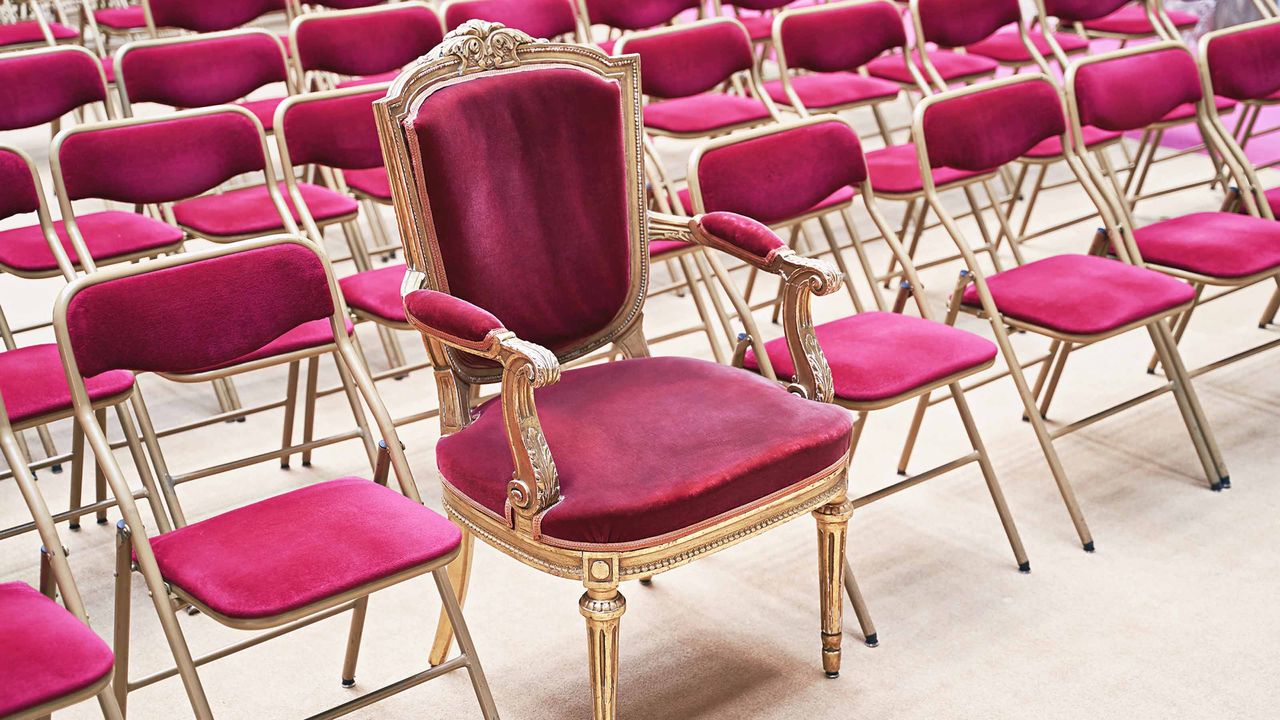 A fancy throne sits in a line of folding chairs.