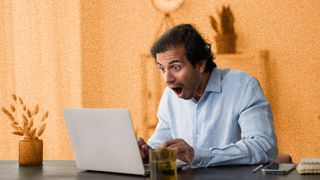 Young man looking shocked, sitting at table looking at laptop.