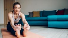 a woman doing a forward fold with fingers wrapped around the back of her feet, facing the camera in a living room setting with a turquoise sofa behind her.