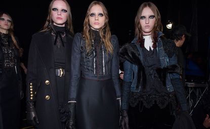 Four female models wearing looks from the Dsquared2 collection. One model is wearing a black and lace piece and black jacket. Another model is wearing a black high neck top with a bow, belt and black jacket with gold buttons. The third model is wearing a blue denim jacket with black detail and a dark blue skirt. And the fourth model is wearing a black aand white piece with lace, a blue denim jacket with black fur and lace and deep red tassel earrings. All the models are wearing black gloves