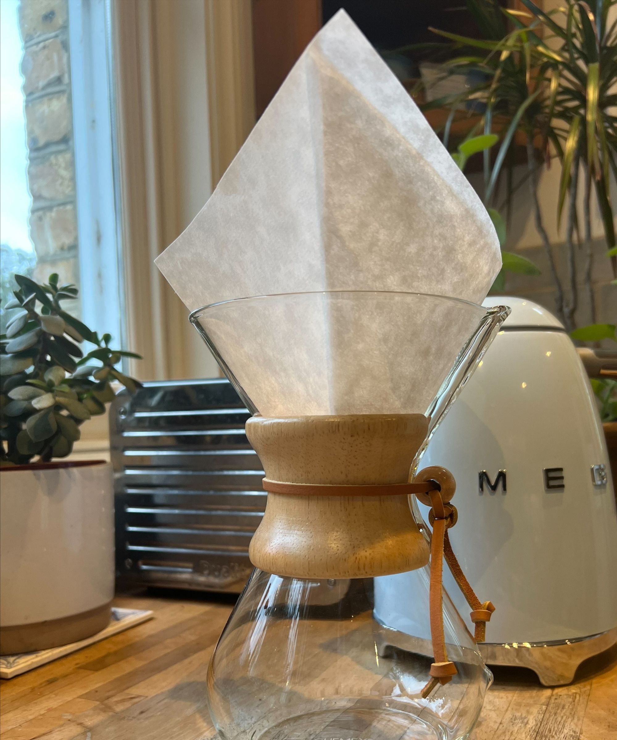A glass CHEMEX coffee maker with a white filter. In the background is a small white pot with a small plant, silver Dualit toaster, baby blue Smeg kettle, and large houseplant with a baby blue backsplash and wooden corner cabinet. To the left is a window, surrounded by yellow brick on the outside.