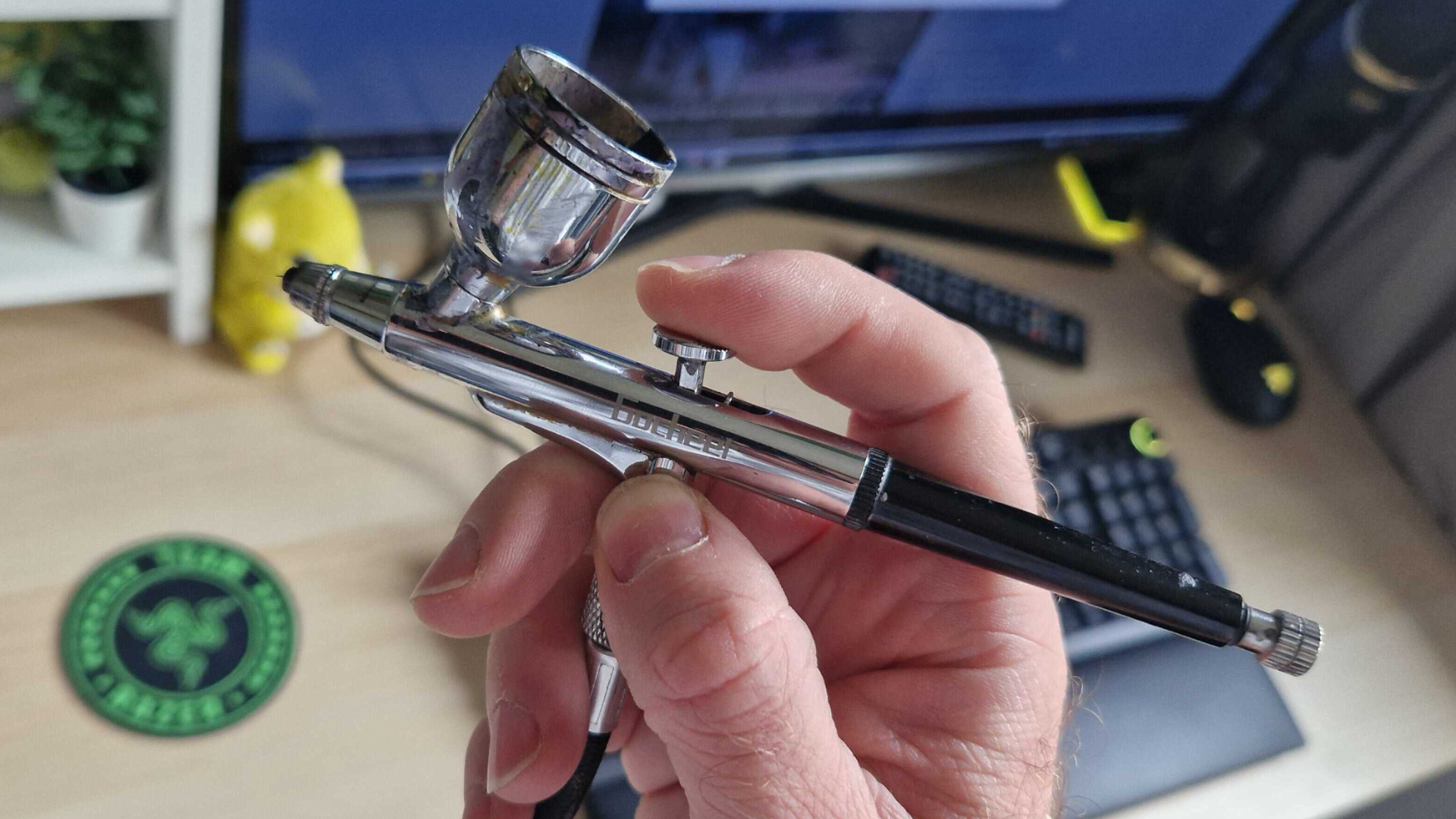 A hand holds up an airbrush in front of a keyboard, mouse, and monitor