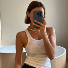 A woman wears a white ribbed tank top with a beaded yellow necklace and black pants.