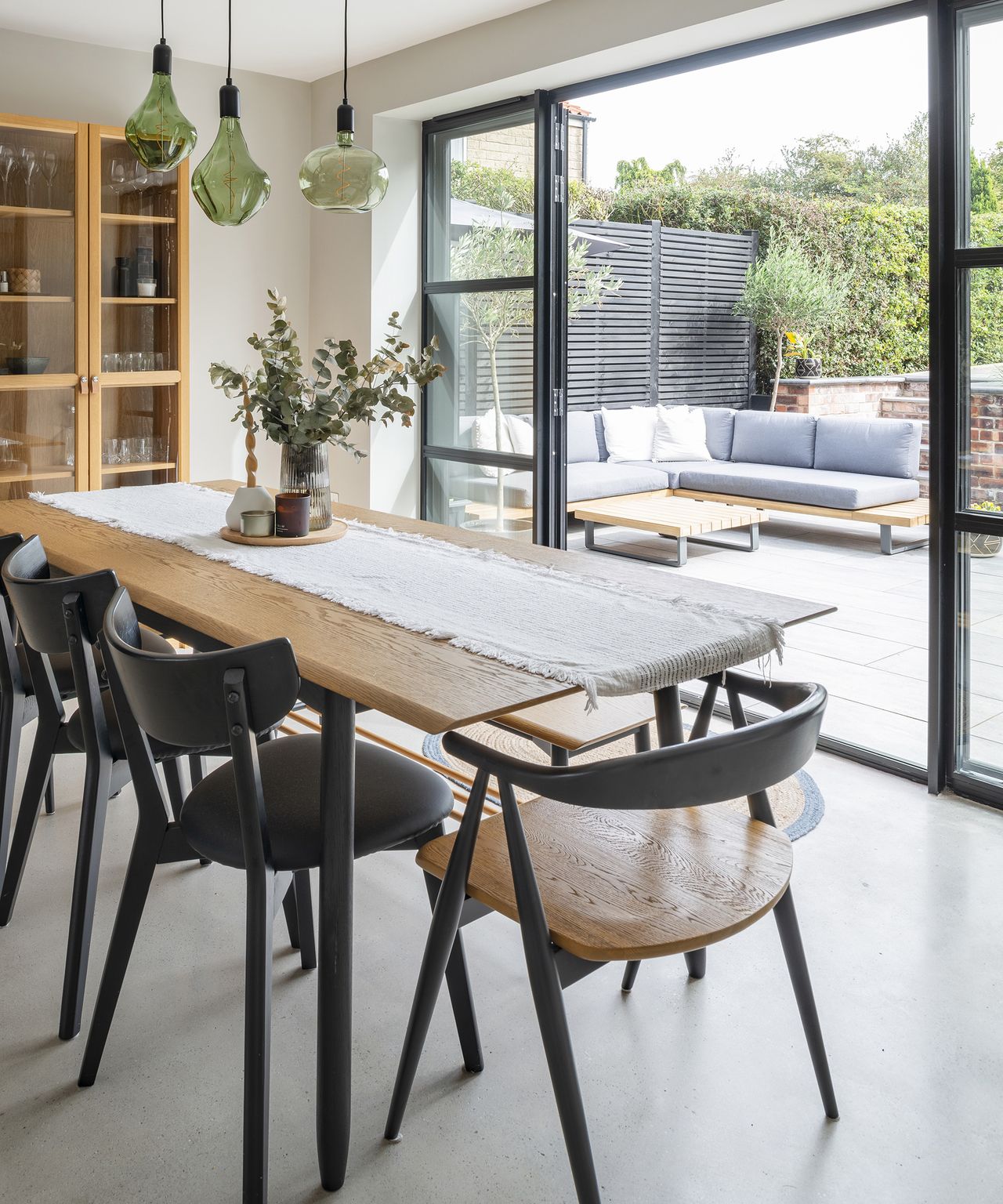 A beautifully styled dining table which looks out through open glass patio doors to a garden. The interior style is Japandi with pale wood and flooring with dark accents