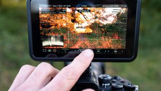 A finger touching the screen of an Atomos Shinobi II camera monitor on top of a camera outdoors in a field