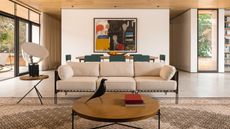 A metal frame sofa with cream cushions in a contemporary living room. A round wooden coffee table in the foreground, and a large dining table with blue chairs around it in the background.