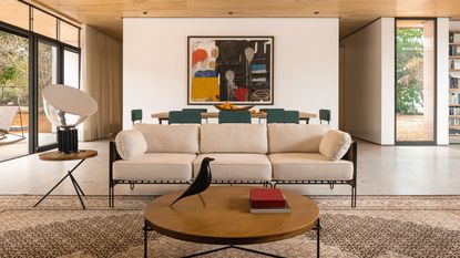A metal frame sofa with cream cushions in a contemporary living room. A round wooden coffee table in the foreground, and a large dining table with blue chairs around it in the background.