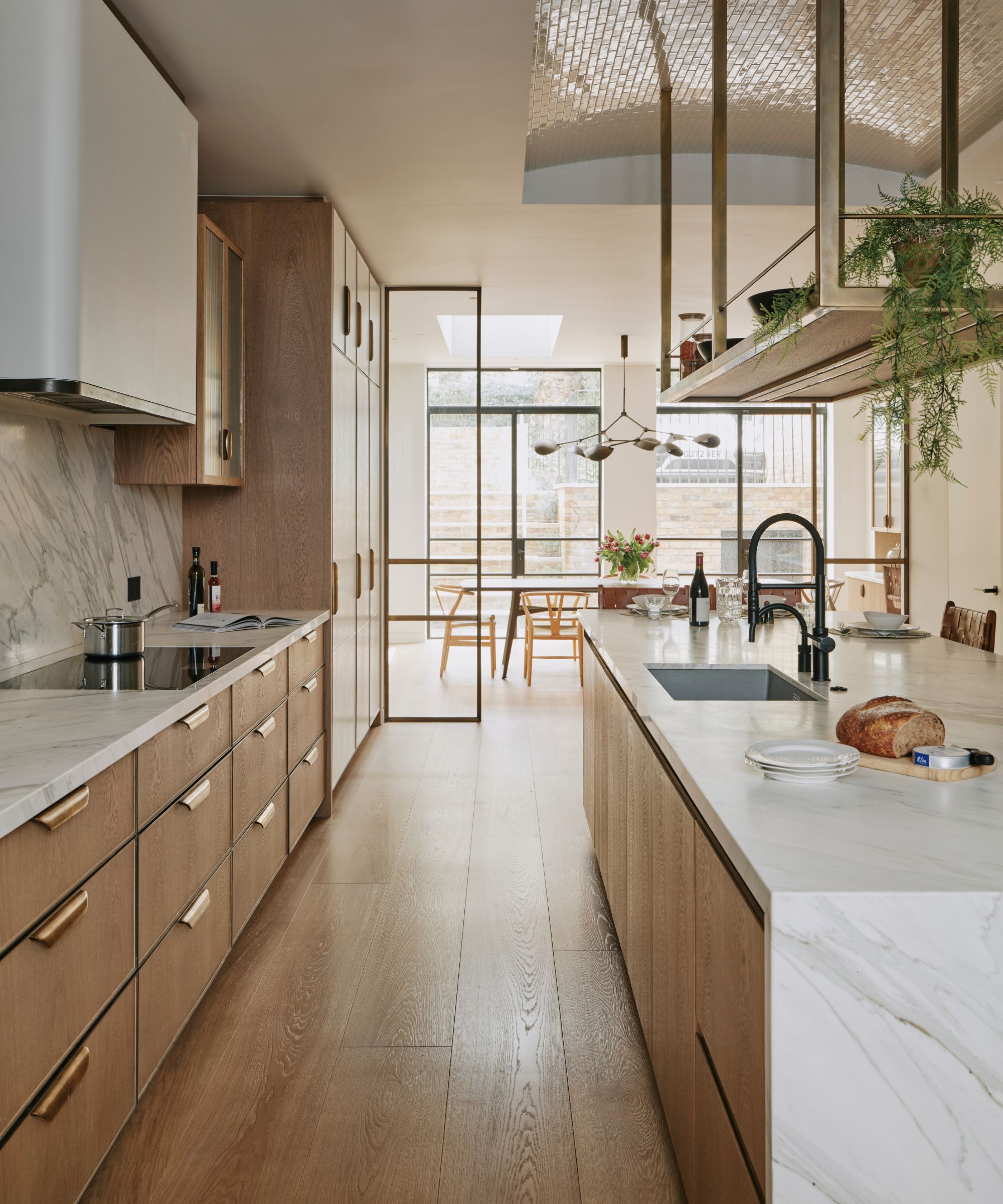 A wooden kitchen with metallic handles and a large marble island overlooks a dining room