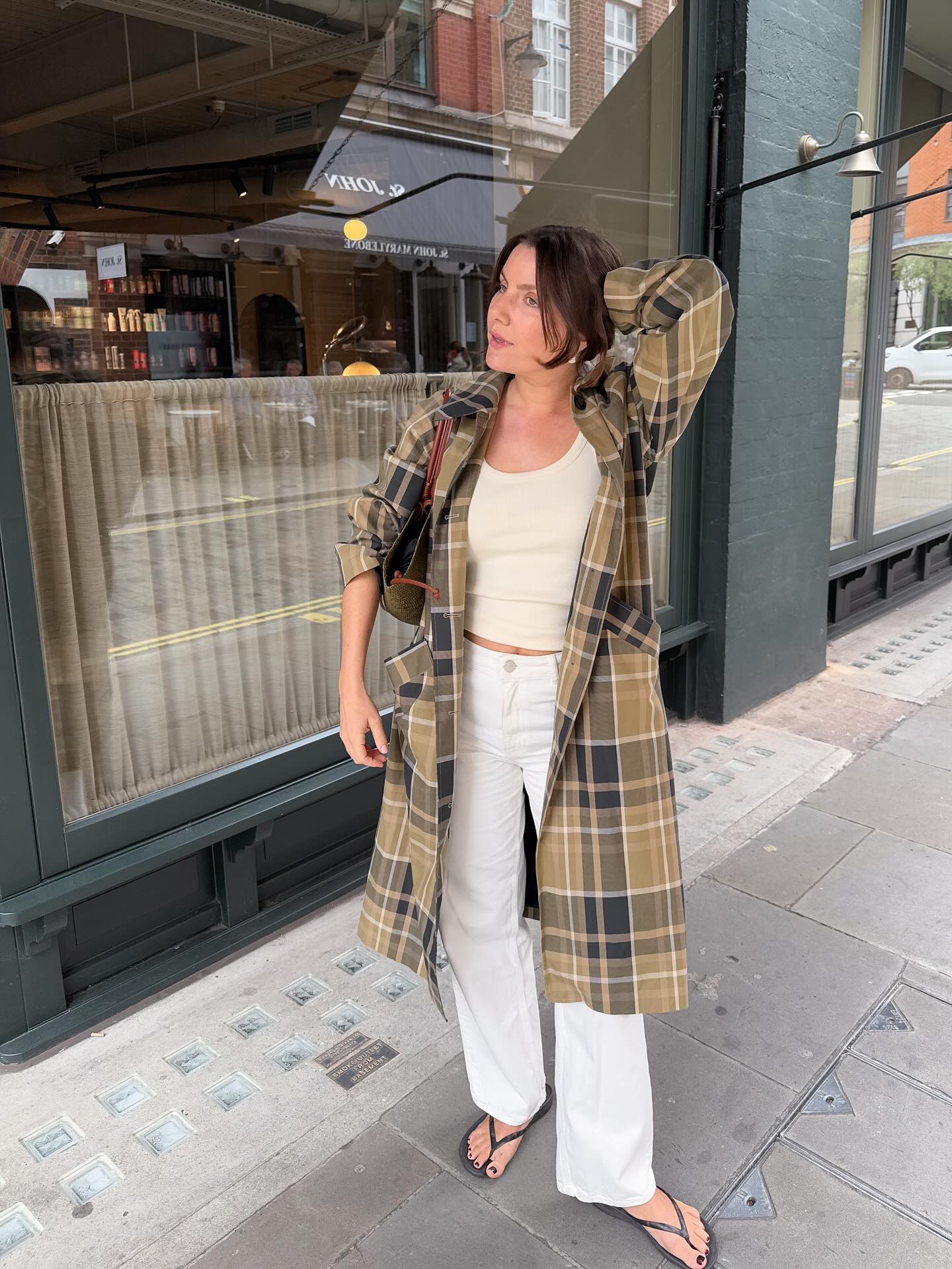 French female fashion influencer Emma Hoareau poses on a London sidewalk wearing a Sezane plaid jacket, a basic tank top, white jeans, and black flip flop sandals