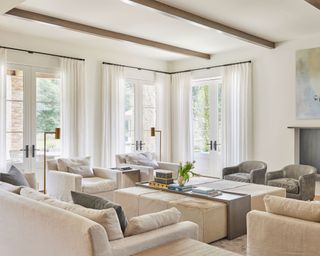 Large living room with sleek dark wooden ceiling beams, white painted walls, large seating area with cream sofas and armchairs, two gray smaller armchairs, large ottoman with tray of decorative objects and accessories