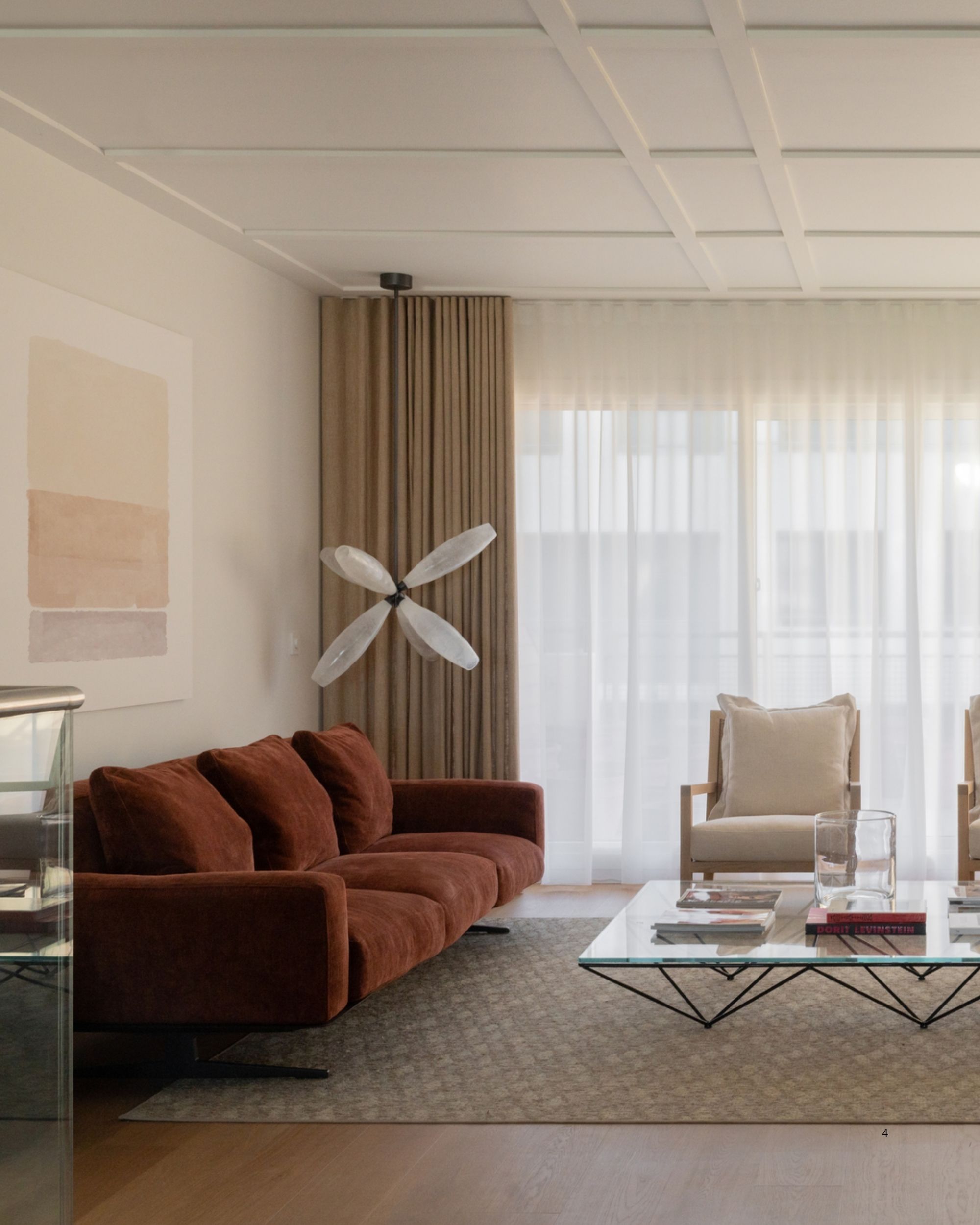 A white and beige living room with curtains over the back window wall, a red sofa, white accent chairs, a pink side chair, and a large glass coffee table.